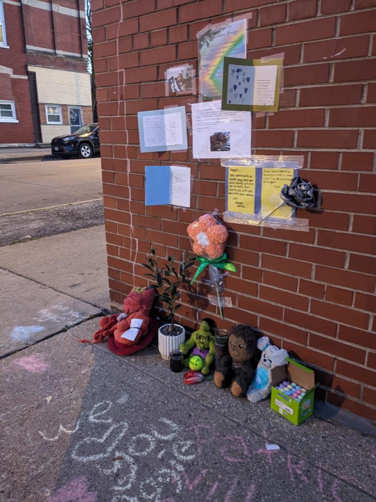 Cards, flowers, toys, candles, and chalk messages arranged beside a brick wall as a memorial for Aggie